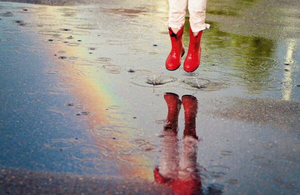 Eine Person springt in eine Pfütze mit „Ich singe im Regen – … und Gummistiefel liegen sowieso im Trend …“.
