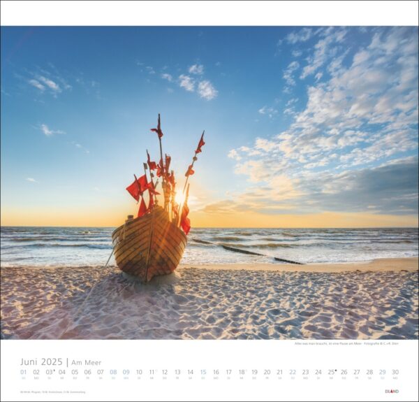 Ein dekoratives Holzboot mit roten Segeln an einem Sandstrand bei Sonnenuntergang, mit Blick auf das Meer unter einem klaren Himmel. Das Boot ist Teil eines Am Meer 2025-Kalenders mit angezeigten Daten.