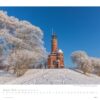 Eine Winterlandschaft zeigt einen historischen Leuchtturm aus rotem Backstein in einer schneebedeckten Landschaft. Raureif auf den Bäumen unter einem klaren blauen Himmel, mit einem Fußweg, der zur Faszination Leuchttürme – An den Grenzen der Landschaft 2025 führt.