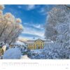 Eine Winterlandschaft in Schleswig-Holstein 2025 zeigt ein großes gelbes Gebäude mit weißen Verzierungen, eingebettet zwischen schneebedeckten Bäumen unter einem strahlend blauen Himmel. Die Szene, gerahmt als Kalender für Februar