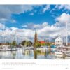 Ein Kalenderblatt für Schleswig-Holstein 2025 mit einer malerischen Szene aus Schleswig-Holstein mit einem Yachthafen voller Segelboote im Vordergrund und einer markanten Kirche mit einem hohen grünen Turm darunter.