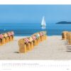 Eine ruhige Strandszene mit einer Reihe nummerierter Strandkörbe mit Blick auf das Meer und einem Segelboot in der Ferne unter einem klaren blauen Himmel. Unten ist ein Kalender für Schleswig-Holstein 2025 eingeblendet.