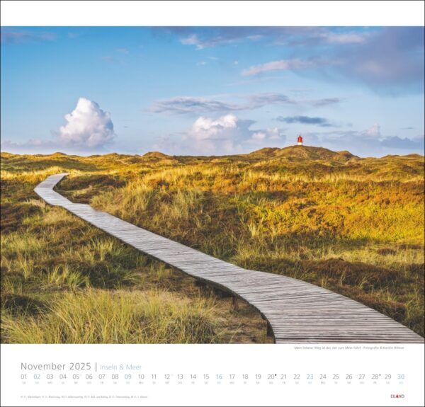Ein Inseln & Meer-Kalender 2025 mit einer ruhigen Landschaft mit einem gewundenen Holzweg durch eine grasbewachsene Düne unter einem blauen Himmel mit flauschigen Wolken. Eine entfernte Figur in Rot steht auf einer entfernten Düne