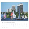 Ein lebendiges Foto, das ein Segelbootrennen an einem sonnigen Tag in der Hansestadt Hamburg zeigt – PostkartenKalender 2025. Es zeigt sieben Segelboote mit bunten Segeln auf dem Wasser vor der Kulisse moderner, hoher Gebäude.