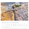 Eine kreative Sandskulptur einer Meerjungfrau mit grünem Seetanghaar und einer Muschelkette liegt an einem Strand, abgebildet auf einer Kalenderseite „Watt’n Meer – PostkartenKalender 2025“ vom Oktober 2025 mit deutschem Text.