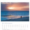 Eine Seite des Nordseeküste - PostkartenKalender 2025 für Dezember zeigt eine ruhige Strandszene bei Sonnenuntergang. Eine einsame Gestalt steht am Wasser und beobachtet einen Vogelschwarm am Ufer. Der Himmel und