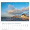 Ein Schleswig-Holstein – PostkartenKalender 2025 mit einem ruhigen Strandbild, das Häuser entlang der Küste unter einem klaren Himmel während des Sonnenuntergangs zeigt.