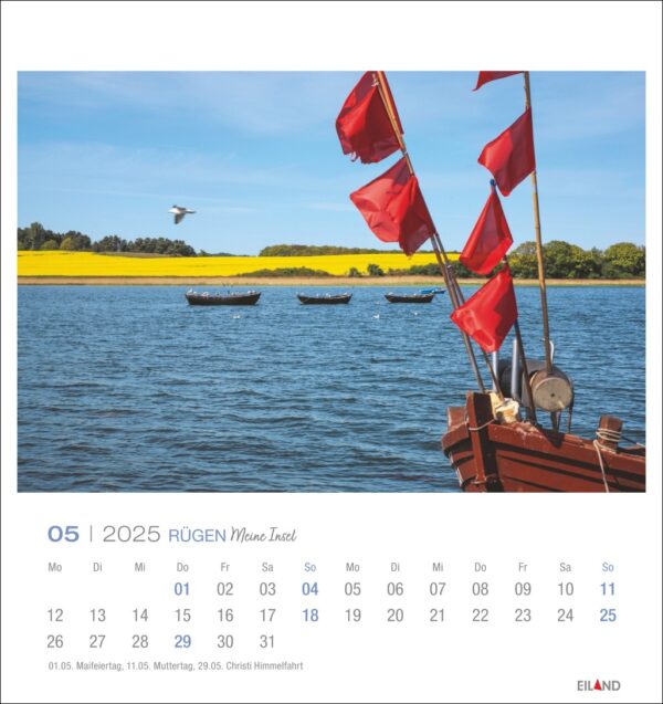 Eine Rügen - PostkartenKalender 2025 Seite für den Mai mit einer heiteren Seenlandschaft: rote Fahnen flattern auf einem Boot im Vordergrund, klarer blauer Himmel, leuchtend grüne Bäume im Hintergrund, kleine Boote