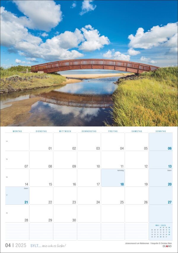 Ein Kalenderblatt für Sylt … wie ich es liebe 2025 mit einem großen Landschaftsfoto, das eine Holzbrücke zeigt, die sich über einen spiegelnden Fluss wölbt, vor der Kulisse eines strahlend blauen Himmels mit flauschigen Wolken.
