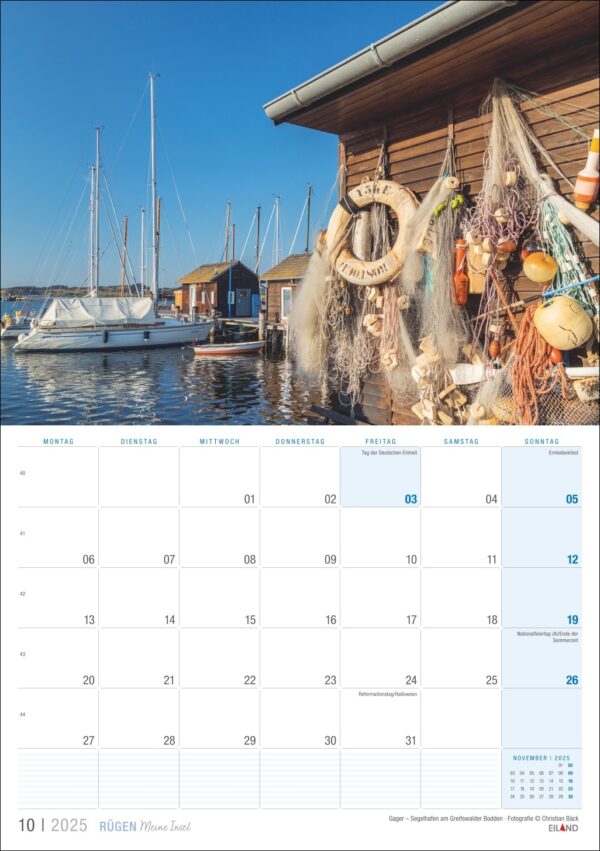 Eine Kalenderseite für Rügen …meine Insel 2025 zeigt einen ruhigen Yachthafen auf der Insel Rügen mit Yachten, die in der Nähe einer mit nautischem Dekor geschmückten Holzhütte angedockt sind. Der obere Teil der Seite