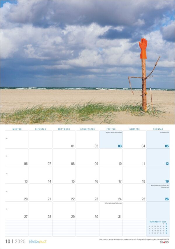 Ein Bild einer Waterkant-Kalenderseite für Oktober 2025, die eine surreale Waterkant-Strandszene mit einem Holzpfosten zeigt, auf dessen Spitze ein orangefarbener Handschuh nach oben zeigt, vor einem bewölkten Himmel und mit Gras gesäumt.