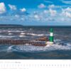 Ein „Küstenland – Meer erleben“-Kalender 2025 zeigt einen Leuchtturm mit grüner Spitze auf einem Wellenbrecher inmitten einer rauen See unter einem wolkenverhangenen blauen Himmel, mit aufgeführten Tagen und Daten.