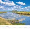Ein malerischer Küstenland - Meer erleben-Kalender 2025 mit einer Landschaft mit einem Schloss auf einem entfernten Hügel, umgeben von üppigem Grün und Sümpfen. Eine kleine hölzerne Fußgängerbrücke überquert das nahe Wasser unter einem blauen Himmel.