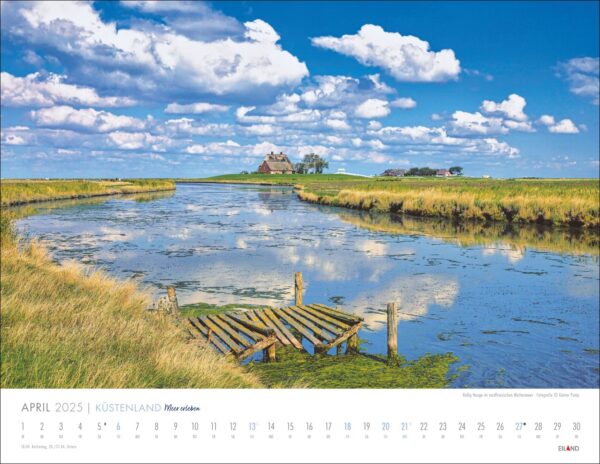 Ein malerischer Küstenland - Meer erleben-Kalender 2025 mit einer Landschaft mit einem Schloss auf einem entfernten Hügel, umgeben von üppigem Grün und Sümpfen. Eine kleine hölzerne Fußgängerbrücke überquert das nahe Wasser unter einem blauen Himmel.