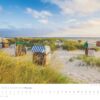 Ein Küstenland - Meer erleben 2025 August Kalenderbild mit einer ruhigen Strandszene in Küstenland, mit verstreuten Strandkörben auf Sanddünen. Sanfte Wellen vom Meer erleben treffen das Ufer unter einem