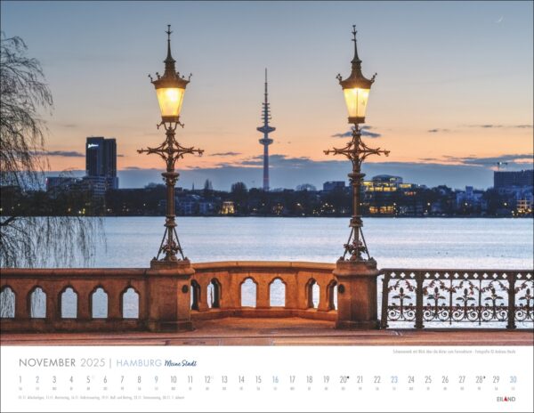 Eine malerische Aussicht auf Hamburg - Meine Stadt 2025 mit einer von Lampen beleuchteten Steinbrücke über einem ruhigen See vor einem Abendhimmel. Im Hintergrund der Skyline der Stadt ist die markante Silhouette eines Fernsehturms zu sehen. Das Bild wird verwendet