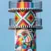 Ein vertikaler LongEiland - Kalender 2025 für Juli mit einem Leuchtturm, der mit einem komplizierten, farbenfrohen Mosaikmuster verziert ist. Die Kalenderdaten sind rechtsbündig ausgerichtet, mit deutschen Abkürzungen für die Monate.