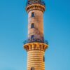 Vertikales LilleEiland - Kalender 2025-Layout für März mit einem Leuchtturm in Warnemünde, Deutschland. Der Turm steht vor einem blauen Himmel, ist mit gelben und braunen Ziegelmustern und einem markanten runden Balkon verziert.