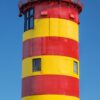 Ein farbenfroher Leuchtturm mit roten und gelben Streifen unter einem klaren blauen Himmel auf einer „LilleEiland – Kalender 2025“-Seite, einschließlich deutschem Text und Daten vom 1. bis 31.