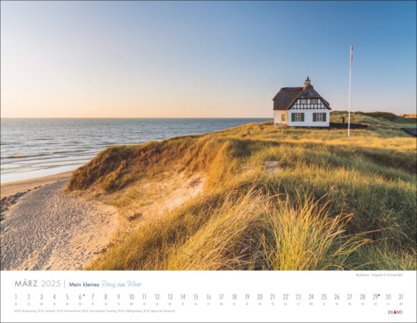 Eine heitere Szene von „Mein kleines Haus am Meer 2025“ auf einem Sandhügel mit windgepeitschten Dünen, mit Blick auf einen ruhigen Strand bei Sonnenuntergang.