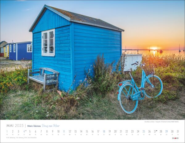 Ein leuchtend blaues „Mein kleines Haus am Meer“ und ein passendes Fahrrad auf grasbewachsenem Küstenland bei Sonnenuntergang. Ein Kalender für Mai 2025 überlagert den unteren Teil des Bildes, wobei die Tage deutlich markiert sind. Die Atmosphäre ist heiter