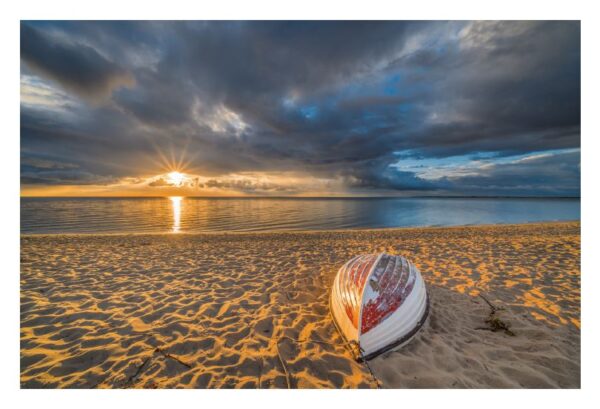 Ein lebhafter Sonnenuntergang über einem Strand auf meiner Insel FÖHR mit bauschigen Wolken am Himmel und der Sonne, die goldenes Licht spendet. Ein umgekipptes rot-weißes Boot liegt auf dem strukturierten Sand im Vordergrund.