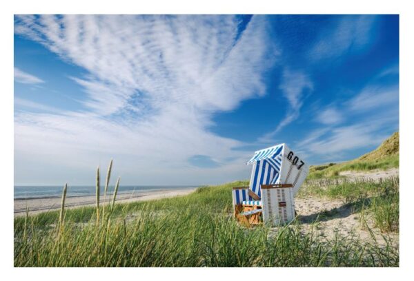 Ein gestreifter Strandstuhl steht auf einer sandigen BeachBumMat mit Blick aufs Meer, umgeben von üppigem grünem Gras und Dünen unter einem strahlend blauen Himmel mit dünnen Wolken.