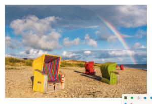 Drei bunte Strandstühle – gelb mit Streifen, rot und grün – stehen mit Blick auf das Meer an einem Sandstrand auf der Insel Föhr unter einem blauen Himmel mit flauschigen Wolken und einem leuchtenden Regenbogen im Hintergrund.