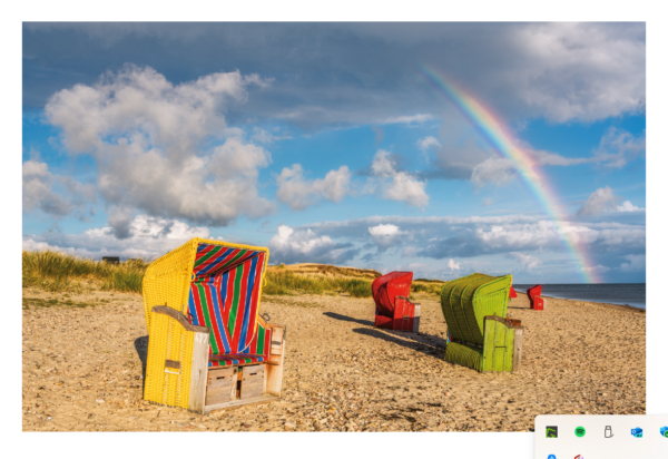 Drei bunte Strandstühle – gelb mit Streifen, rot und grün – stehen mit Blick auf das Meer an einem Sandstrand auf der Insel Föhr unter einem blauen Himmel mit flauschigen Wolken und einem leuchtenden Regenbogen im Hintergrund.