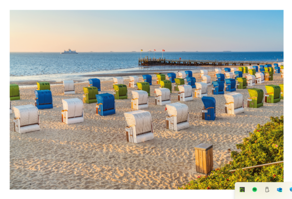 Bunte FÖHR ...meine Insel-Stühle säumen einen Sandstrand in der Nähe einer Anlegestelle auf der Insel Föhr, mit dem Meer und einem Schiff am Horizont, unter einem klaren blauen Himmel während des Sonnenuntergangs.