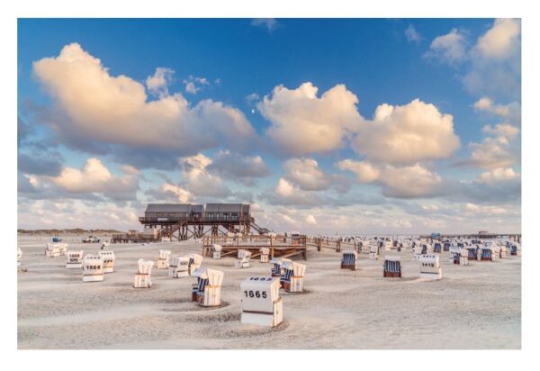 Ein Sandstrand mit zahlreichen weiß-blau gestreiften Küstenland-Liegestühlen mit Blick auf das Meer, unter einem Himmel voller flauschiger Wolken bei Sonnenuntergang. Im Hintergrund steht ein großes, auf Stelzen stehendes Holzgebäude.