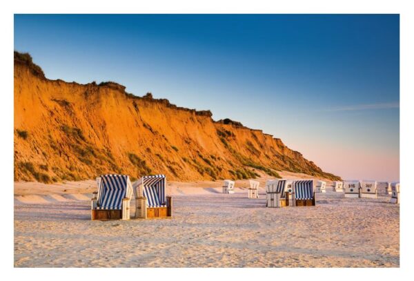 Gestreifte Strandstühle blicken bei Sonnenuntergang auf das Meer. Sie stehen auf Sandstränden unterhalb einer hoch aufragenden, orangefarbenen Klippe auf der ruhigen Insel Sylt und vermitteln eine ruhige Küstenatmosphäre.