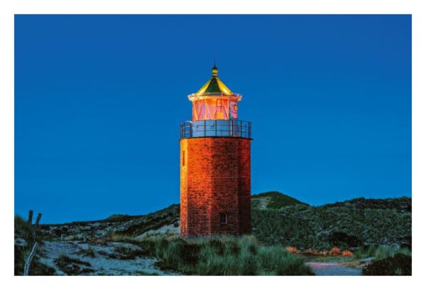 Ein beleuchteter Leuchtturm aus Ziegelsteinen steht vor einem Abendhimmel, sein Licht strahlt warm auf die Sylter Insel. Das Bauwerk liegt zwischen Dünen mit spärlicher Vegetation und unterstreicht seine Abgeschiedenheit und die Ruhe