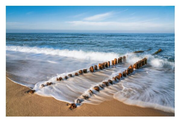 Eine ruhige Strandszene auf „Sylt ...meine Insel“ mit alten Holzpfählen, die aus dem Sand ragen und ins Meer führen. Sanfte Wellen in Bewegung erzeugen eine weiße, schäumende Brandung entlang der Küste vor einem