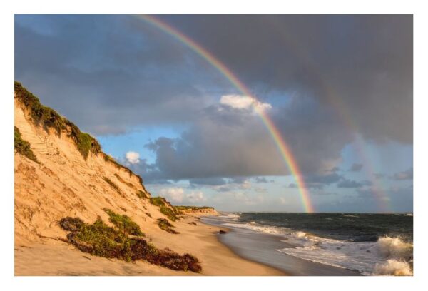 Ein doppelter Regenbogen wölbt sich über dem Sandstrand von Sylt ...meine Insel mit einer steilen, mit Vegetation bedeckten Düne. Goldenes Sonnenlicht erhellt die Szene und hebt die leuchtenden Farben des Regenbogens und die raue Textur hervor.