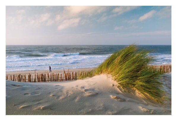 Eine ruhige Strandszene auf Sylt ...meine Insel mit einer Person, die am Ufer entlang geht. Im Vordergrund ist eine große Sanddüne mit hohem Strandhafer zu sehen, der sich im Wind biegt und von einem Holzzaun begrenzt wird.