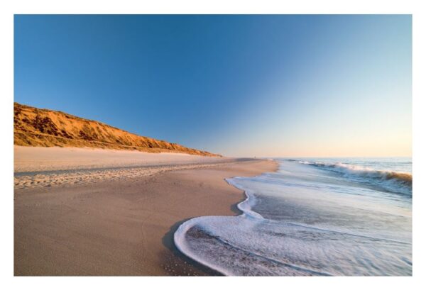 Eine ruhige Strandszene bei Sonnenuntergang auf Sylt ...meine Insel, mit sanften Wellen, die an Sandstrände schlagen. Die linke Seite zeigt eine abfallende Sanddüne unter einem klaren, abfallenden Himmel, der von