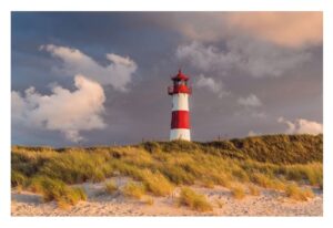 Ein rot-weißer Leuchtturm steht hinter einem Sandstrand mit Büscheln aus hohem Gras auf Sylt ...meine Insel, unter einem Himmel voller goldenem Sonnenlicht und dramatischen Wolken.
