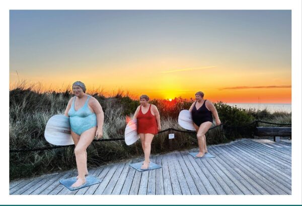 Drei Frauenfiguren, jede mit einem Surfbrett in der Hand, stehen bei Sonnenuntergang auf einem Holzsteg am Strand von SYLT ...meine Insel. Der Himmel ist in leuchtenden Orange- und Blautönen gehalten.